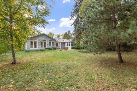 A light gray house with a green lawn and surrounded by trees in the backyard.