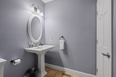 Minimalistic bathroom with a lavender wall, white pedestal sink, and round mirror with fixtures.
