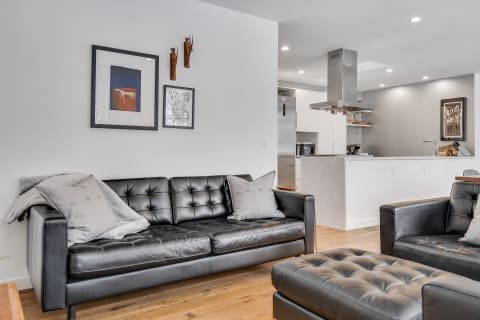 Modern living room featuring a black leather couch, framed artworks, and a minimalistic kitchen in the background.