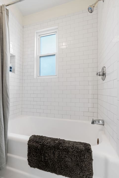 A bright bathroom with white subway tiles and a dark bath mat on the tub edge.