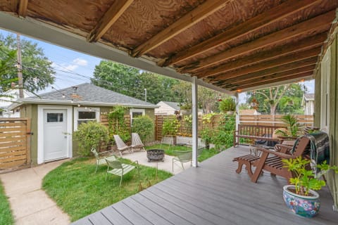A view of a backyard featuring a covered porch, a fire pit, and a shed, surrounded by greenery.