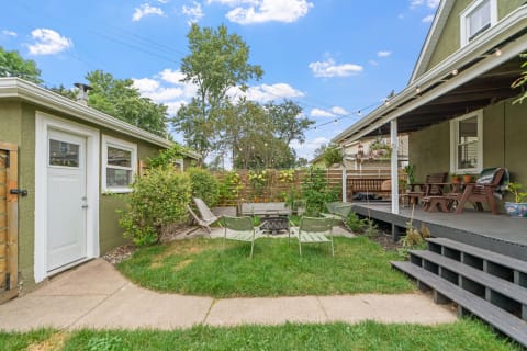 A sunny backyard featuring a fire pit surrounded by light green chairs, lush grass, and a wooden deck.