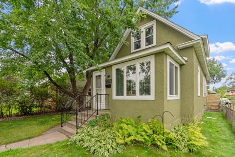 A cozy green house surrounded by greenery and trees under a blue sky.