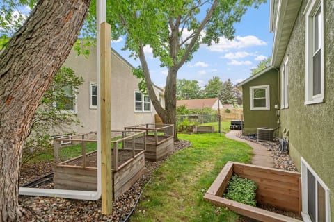 A side yard view featuring garden beds, a large tree, and a green house.