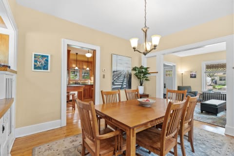 A bright dining room with a wooden table, chandelier, and a connecting kitchen area.