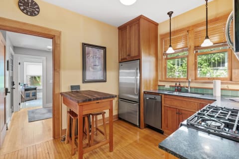A bright kitchen with wooden cabinets, a stainless steel refrigerator, a small table, and sunlight streaming through the windows.