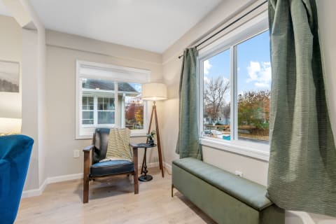 A corner of a room with a black armchair, side table, floor lamp, and a view of colorful trees outside the window