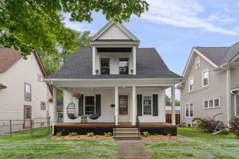 A white two-story house with a front porch and hanging chairs surrounded by greenery.