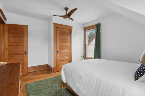A neatly arranged bedroom with a white bedspread, wooden doors, and a window with green curtains.