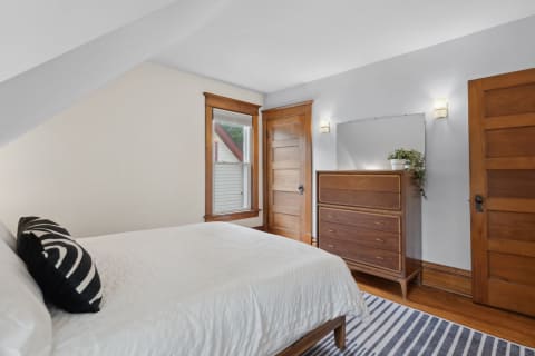 Cozy bedroom featuring a white bedspread, black and white pillow, wooden dresser, and natural light from a window.