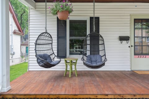 Front porch featuring two hanging wicker chairs, a green stool, and a potted flower plant.