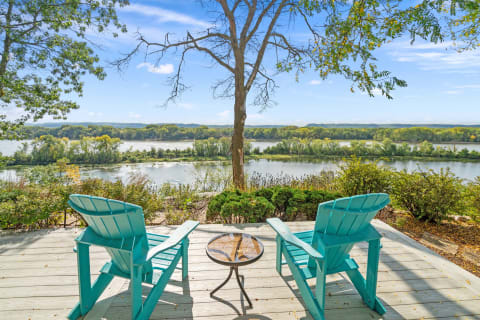 Two turquoise Adirondack chairs on a wooden deck overlooking a peaceful river with trees in autumn colors.