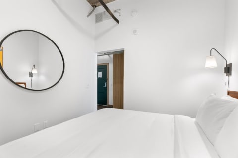 A minimalist bedroom with a round mirror, wall lamp, and neatly made bed.