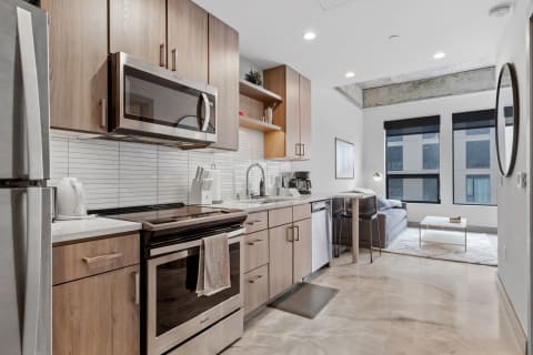 Interior of a modern kitchen with light wooden cabinets and a living area featuring a gray couch and large windows.