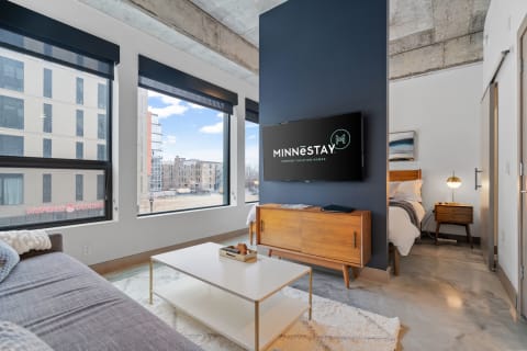 Interior of a contemporary living room with a sofa, coffee table, andmounted TV, showcasing large windows with city views.