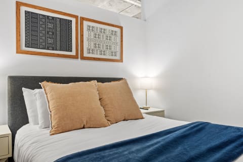 A cozy bedroom featuring a gray upholstered bed, large decorative pillows, blue throw, and framed artwork on the wall.