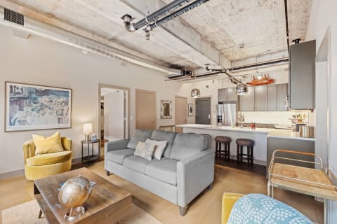 A modern apartment interior featuring a grey sofa, yellow chair, wooden coffee table, and a kitchen backdrop with sleek cabinetry.