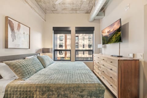 Minimalist bedroom with green bedding, abstract art, and large windows overlooking an urban view.