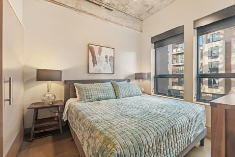 A modern bedroom with teal bedding, bedside lamps, and large windows showcasing an urban view.