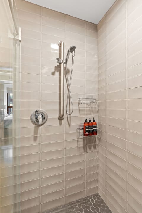 Contemporary shower featuring beige tiles, a chrome faucet, and shower products on an acrylic shelf.