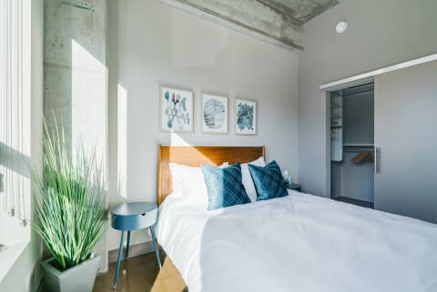 A modern bedroom with a cozy bed, teal decorative pillows, and a plant, illuminated by natural light.