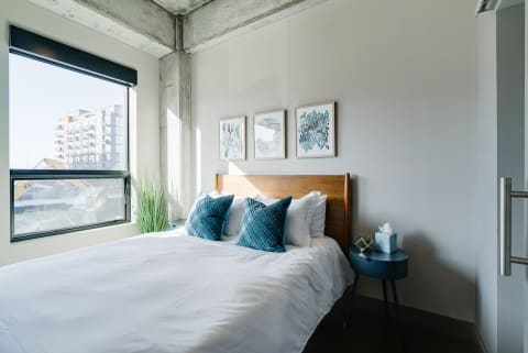 A minimalist bedroom featuring a bed with white linens and blue pillows, framed art on the wall, and a side table with a gift and tissue box.