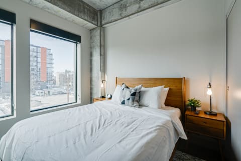 A bright and airy bedroom featuring a wooden bed, geometric pillows, and large windows overlooking an urban landscape.