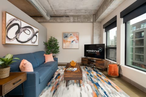 Modern living room with blue sofa, abstract art, and a television displaying MINNESTAY logo.