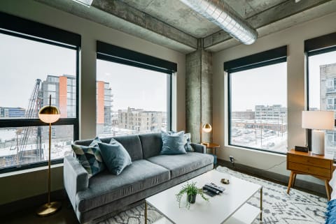 Modern living room featuring a gray sofa, blue pillows, large windows, and an urban skyline.