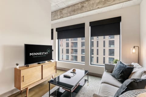 A contemporary living room featuring a gray couch, black coffee table, and television with MINNĒSTAY logo.