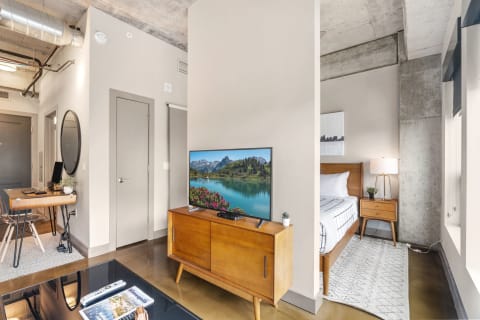 A contemporary living space with a wooden TV stand, desk area, and a well-made bed.