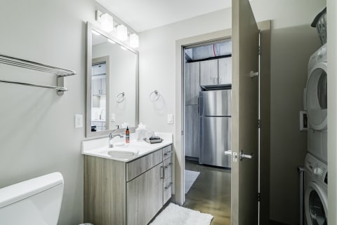 Contemporary bathroom with light gray walls, a wooden vanity, and a doorway leading to a kitchen.