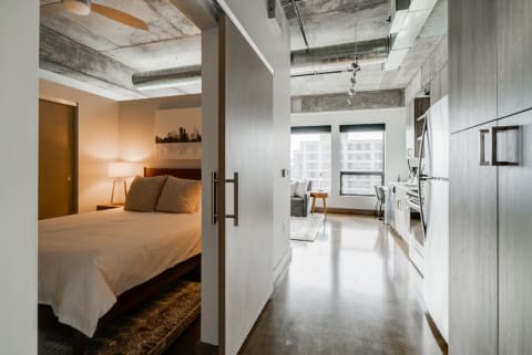View of a modern studio apartment featuring a bedroom, kitchen, and large windows.