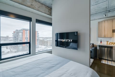 A spacious bedroom featuring a bed, a wall-mounted TV displaying 'MINNESTAY', and large windows showing a snowy cityscape.