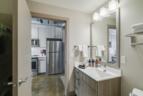 Interior view of a modern bathroom leading to a kitchen, showcasing a sink, toiletries, and contemporary styling.