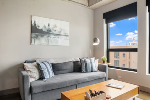 A living room featuring a gray sofa with blue and white striped pillows, modern lamp, and a large window with an urban view.