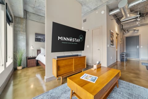 Modern living room with a large TV displaying MINNēSTAY logo, stylish furniture, and polished concrete floors.