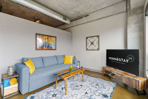 A cozy living room featuring a blue sofa, yellow cushions, art on the wall, and a television displaying MINNESTAY logo.