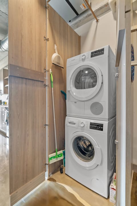 A stacked washer and dryer unit in a wooden-paneled laundry space with cleaning tools.