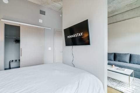 Modern bedroom with a television showing 'MINNESTAY', a cozy bed, and a gray sofa.