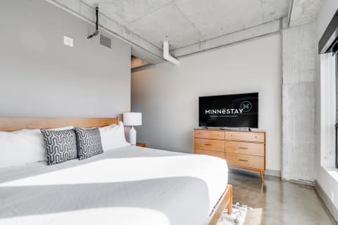 A minimalist bedroom with a wooden bed, gray pillows, a TV, and polished concrete floor.
