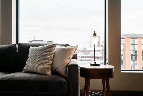 Cozy living room corner with a gray couch and a lamp, featuring a city view through large windows.