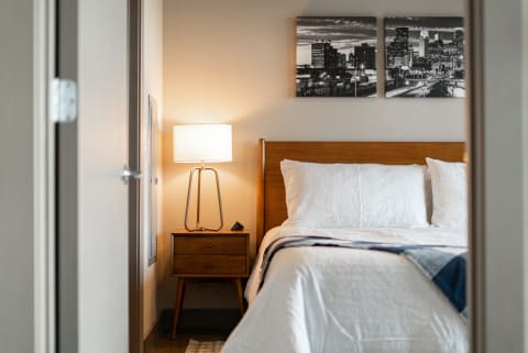 A cozy modern bedroom with white bedding and a stylish lamp, viewed through an open door.