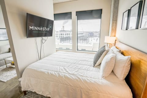 A cozy bedroom featuring a modern bed, a TV on the wall, and large windows.
