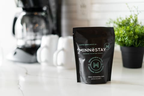 A black coffee bag from MINNESTAY beside white mugs and a coffee machine on a marble countertop.