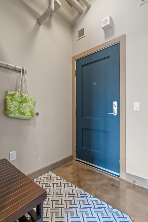 A minimalist entryway with a blue front door, dark wooden bench, green bag, and patterned rug.
