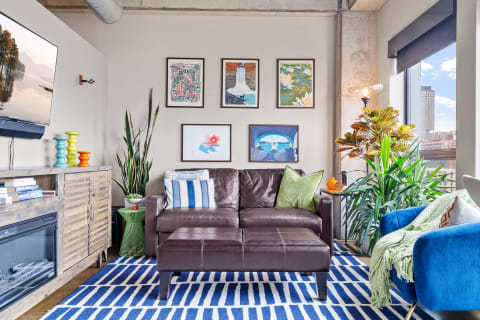 A cozy living room with a brown leather sofa, colorful artwork on the walls, and green plants.