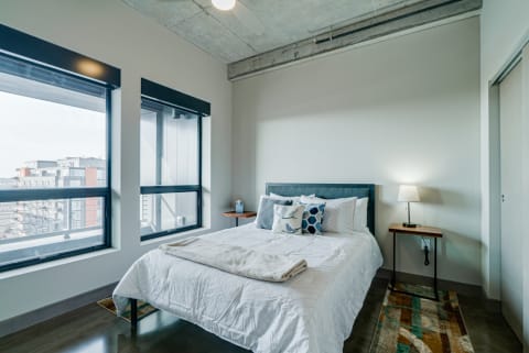 Modern bedroom featuring a blue upholstered bed, decorative pillows, and large windows with city views.