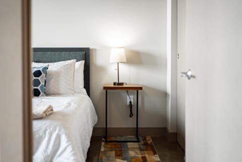 A minimalistic bedroom featuring a bed, nightstand with a lamp, and a colorful rug.