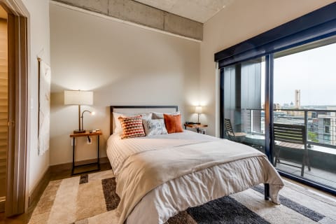 A stylish bedroom featuring a bed with decorative pillows, a nightstand with a lamp, and large windows overlooking the city.